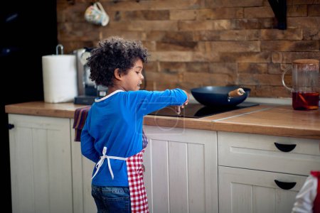 Photo for Beautiful young boy standing in a domestic kitchen by the stove, turning it on, preparing to make a meal. Young chef. Home, family, lifestyle concept. - Royalty Free Image