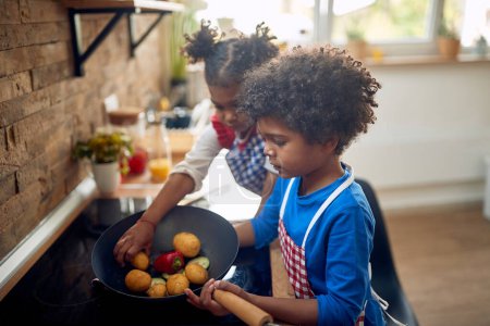 Foto de Dos niños pequeños, hermano y hermana juntos en una cocina doméstica preparando el almuerzo con verduras por la estufa, cocinar en una sartén. Hogar, familia, estilo de vida. - Imagen libre de derechos