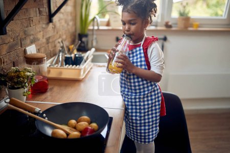 Photo for Cute little girl standing in a kitchen by the stove drinking a juice while the veggies are frying in a pan. Home, family, lifestyle concept. - Royalty Free Image