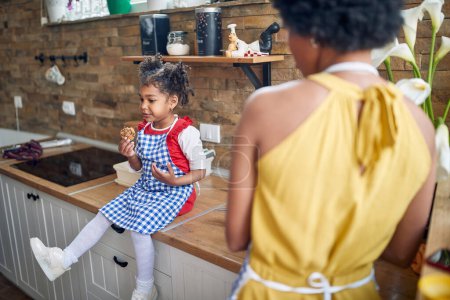 Photo for Afro-American girl sits on the kitchen counter, delightfully enjoying a cookie. Her mother stands by the sink, her gaze filled with love and admiration as she watches her daughter. - Royalty Free Image