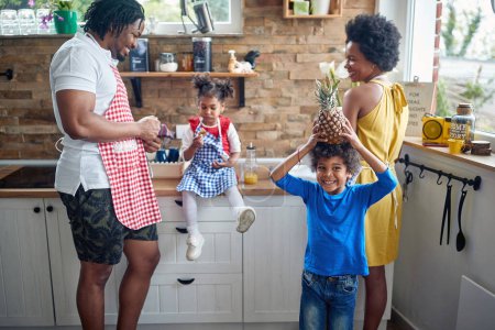 Photo for Beautiful joyful family in a domestic kitchen together, little boy holding pineapple over his head like a crown. Home, family, lifestyle concept. - Royalty Free Image