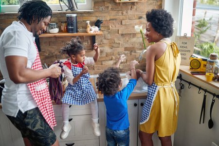 Photo for Afro-American family in the kitchen.The little girl sits on the kitchen counter, delightfully indulging in a delicious treat, while her mother diligently washes the dishes with help by son and father. - Royalty Free Image