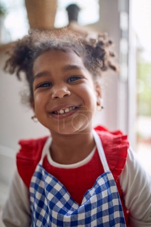 Photo for Cute little young girl child standing in a kitchen waring a blue chekered apron and feeling joyful, smiling. Home, family lifestyle concept. - Royalty Free Image