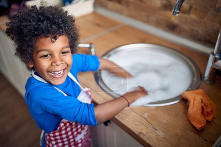 Téléchargez les photos : Le jeune garçon afro-américain prend la responsabilité de laver la vaisselle avec beaucoup d'enthousiasme. Donnant un tablier coloré, il se tient avec confiance à l'évier, entouré d'une mer de mousse savonneuse. - en image libre de droit