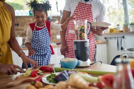 Foto de La familia afroamericana se reúne para preparar una comida en su cocina. La imagen muestra a una madre, padre e hija trabajando en colaboración, rodeados de ingredientes y utensilios culinarios. - Imagen libre de derechos
