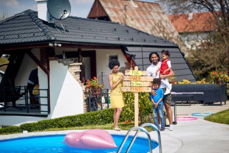 Foto de Familia afroamericana de pie con orgullo al lado de su casa, sosteniendo un cartel que significa la disponibilidad de su casa para la venta o alquiler. - Imagen libre de derechos