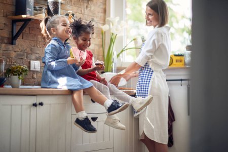 Téléchargez les photos : Deux adorables petites filles assises sur le comptoir de la cuisine mangeant de la pastèque et du muffin ensemble, avec leur mère à côté de leur vaisselle dans une cuisine moderne. Maison, famille, concept de style de vie. - en image libre de droit