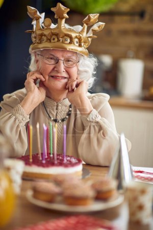 Foto de Una querida abuela se sienta a la mesa, su rostro adornado con una sonrisa radiante que refleja toda una vida de amor y sabiduría. Delante de ella está el pastel de cumpleaños.. - Imagen libre de derechos