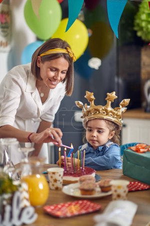 Photo for Lovely birthday girl with golden crown making a wish while mother lights her candles on the cake. Home, family, celebration concept. - Royalty Free Image