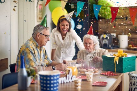 Foto de Celebración de cumpleaños, la abuela soplar las velas en su pastel de cumpleaños.Alrededor de ella no solo son su marido cariñoso, sino también los cuidadores de la casa de cuidado de personas mayores donde reside. - Imagen libre de derechos