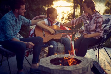 Photo for Young girl playing guitar outdoors by the campfire, mother and father singing along. Lovely family together. Togetherness concept. - Royalty Free Image