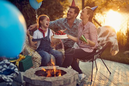 Photo for Joyful happy young birthday girl with cake smeared on her face celebrating birthday with mom and dad, feeling cheerful. Family together in a cottage outdoors. - Royalty Free Image