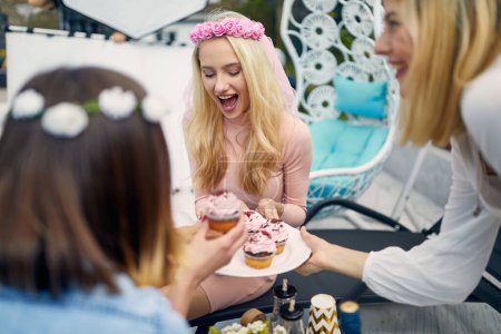 Photo for Young joyful women on a bachelorette party enjoying cupcakes with frosting, sitting outdoors on a sunny day. Celebaration, party, togetherness concept. - Royalty Free Image