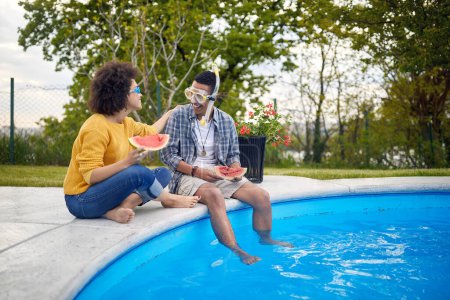 Téléchargez les photos : Sourire beau couple au bord de la piscine avoir du plaisir, et manger de la pastèque - en image libre de droit