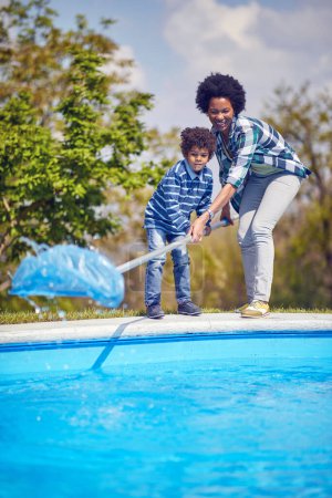 Téléchargez les photos : Jeune mère et son fils nettoyant la piscine avec un filet de nettoyage ensemble debout près de la piscine, travaillant en équipe. Ensemble, mode de vie, concept familial. - en image libre de droit