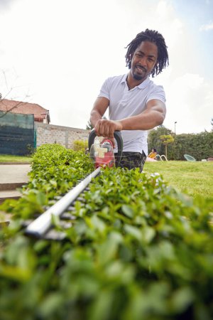 Téléchargez les photos : Jeune homme afro-américain en vêtements décontractés travaillant à l'extérieur dans le jardin à l'aide d'une tondeuse électrique dans la cour arrière. Maison, style de vie, concept de jardinage. - en image libre de droit