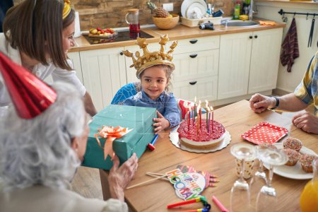 Photo for Happy little girl with an inflatable crown celebrating her birthday, sitting by the table with guests, recieving gifts from her grandmother. Home, family, lifestyle, celebration concept. - Royalty Free Image