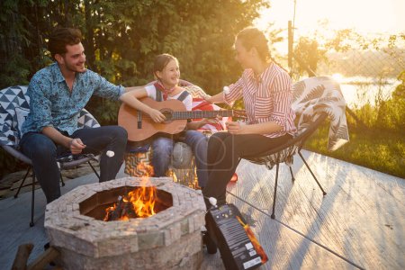 Photo for Happy girl playing guitar outdoors by the fireplace, sitting with mother and father in the backyard enjoying sunny summer day together. Home, family, lifestyle concept. - Royalty Free Image