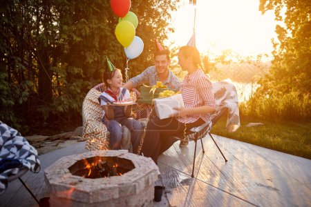 Photo for Young girl celebrating birthday with mother and father, recieving gifts sitting outdoors on a sunny summer day together. Family, celebration, togetherness concept. - Royalty Free Image