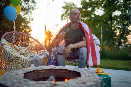 Photo for Horizontal shot of middle aged man sitting outdoors eating marshmallows by a fireplace and celebrating fourth of july. Lifestyle, lesiure, holiday concept. - Royalty Free Image