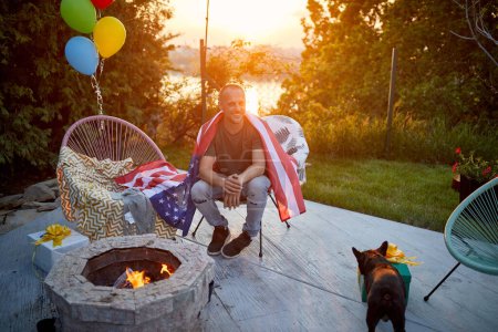 Foto de Alegre hombre de mediana edad con bandera americana alrededor de su espalda, sentado al aire libre junto a una chimenea en su patio trasero, disfrutando de un soleado día de verano con su perro. Estilo de vida, lesión, concepto de hogar. - Imagen libre de derechos