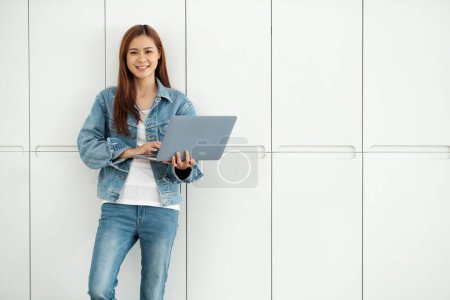 Photo for Young asian female student holding laptop and studying online researching for project with enjoyment while standing agianst wall. Online learning concept. - Royalty Free Image