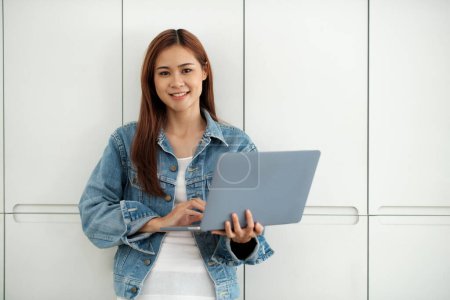 Téléchargez les photos : Jeune étudiante asiatique tenant un ordinateur portable et étudiant la recherche en ligne pour le projet avec plaisir tout en se tenant agianst mur. Concept d'apprentissage en ligne. - en image libre de droit
