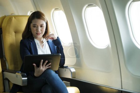 Téléchargez les photos : Jeune femme d'affaires asiatique professionnelle en vêtements formels travaillant à l'aide d'une tablette avec stylo intelligent tout en étant assis dans la cabine de l'avion près de la fenêtre voyageant à un autre endroit. Concept de voyage et d'affaires. - en image libre de droit