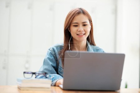 Foto de Jóvenes asiáticos estudiante femenina que estudia en línea con el disfrute de usar el ordenador portátil y el uso de auriculares mientras está sentado en el escritorio con gafas y libros de texto. Concepto de aprendizaje online. - Imagen libre de derechos