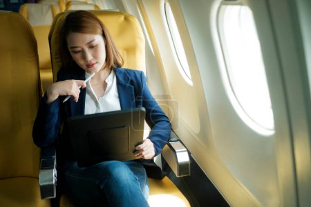 Téléchargez les photos : Jeune femme d'affaires asiatique professionnelle en vêtements formels travaillant à l'aide d'une tablette avec stylo intelligent tout en étant assis dans la cabine de l'avion près de la fenêtre voyageant à un autre endroit. Concept de voyage et d'affaires. - en image libre de droit
