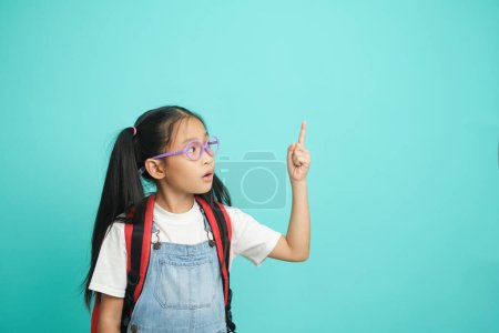 Téléchargez les photos : Gros plan écolière enfant pointant des doigts à l'espace de copie, elle agréable mignon attrayant gai étonné. isolé sur fond bleu pastel. Concept éducatif pour l'école - en image libre de droit