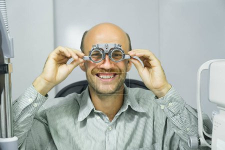 Photo for Smart handsome man sitting in optometrist cabinet having his eyesight checking, examining, testing with trial frame glasses by professional optician for new pairs of eyeglasses. - Royalty Free Image