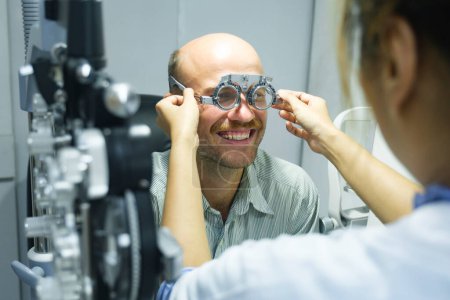 Foto de Hombre guapo inteligente sentado en el gabinete del optometrista que tiene su visión comprobando, examinando, probando con gafas de marco de prueba por óptico profesional para nuevos pares de gafas. - Imagen libre de derechos