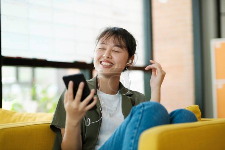 Foto de Mujer asiática joven en auriculares inalámbricos escuchando música usando un teléfono inteligente y relajarse en el sofá en la sala de estar en casa. Conexión online y concepto de aprendizaje en línea. - Imagen libre de derechos