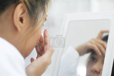 Photo for Young beautiful woman try putting contact lens received from optician presription in her eyes looking at mirror in ophthalmology clinic. - Royalty Free Image