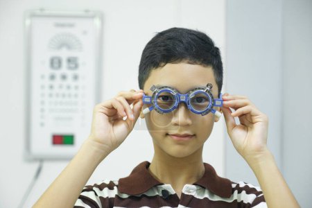 Photo for Smart young boy sitting in optometrist cabinet having his eyesight checking, examining, testing with trial frame glasses by professional optician for new pairs of eyeglasses. - Royalty Free Image