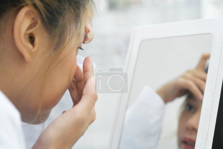 Téléchargez les photos : Jeune belle femme essayer de mettre lentille de contact reçu de la presription opticien dans ses yeux en regardant miroir dans la clinique d'ophtalmologie. - en image libre de droit