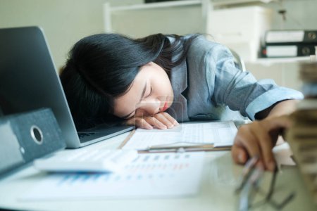 Foto de Joven asiática mujer de negocios mujer cansada durmiendo en el escritorio con el ordenador portátil en el lugar de trabajo solo, siesta en el escritorio delante de su ordenador portátil en la oficina, sobrecargado de trabajo con la cabeza en el escritorio en la oficina. - Imagen libre de derechos