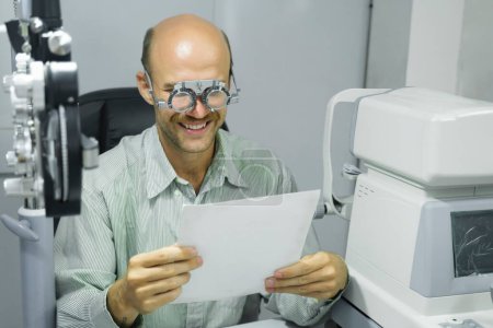 Téléchargez les photos : Charmant homme intelligent assis dans une armoire optométriste ayant sa vue vérifier, examiner, tester avec des lunettes de cadre d'essai par un opticien professionnel pour de nouvelles paires de lunettes. - en image libre de droit