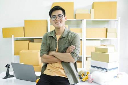 Photo for Young male small business entrepreneur, online store owner working ar workplace or warehouse with shelves of cardboard boxes of customers' order in the background sitting on desk looking cheerfully at camera. Online selling, e-commerce concept. - Royalty Free Image