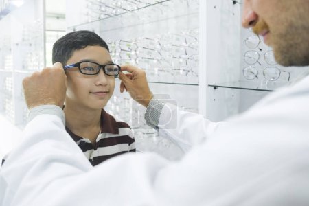 Téléchargez les photos : Smart young acheter étudiant en polo rayé t-shirt heureux d'essayer, tester et choisir de nouvelles lunettes pour l'étude avec opticien professionnel supervisant au magasin d'optique. - en image libre de droit
