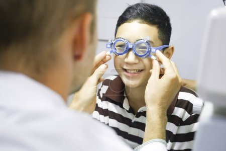 Photo for Smart young boy sitting in optometrist cabinet having his eyesight checking, examining, testing with trial frame glasses by professional optician for new pairs of eyeglasses. - Royalty Free Image