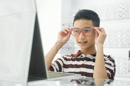 Téléchargez les photos : Smart young acheter étudiant en polo rayé t-shirt heureux d'essayer, tester et choisir de nouvelles lunettes pour l'étude avec opticien professionnel supervisant au magasin d'optique. - en image libre de droit