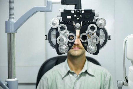 Téléchargez les photos : Charmant homme intelligent ayant un test oculaire, un examen visuel, un contrôle par un opticien professionnel utilisant un phoropter de machine d'essai clinique pour fabriquer de nouvelles lunettes au magasin d'optique. - en image libre de droit