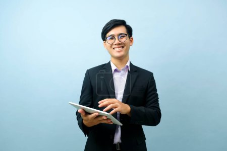 Photo for Portrait of young smart asian businessman wearing glasses standing and smiling while using tablet for working or communicating with light blue isolated background. Business, connection concept. - Royalty Free Image