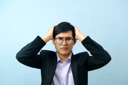 Photo for Portrait of young determined asian businessman wearing eyeglasses standing and putting his hand on his head stressed and frustrated from business work with light blue isolated background. Business concept. - Royalty Free Image