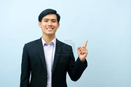 Foto de Portriat of asian young smart happy businessman dressed in suit standing straight, smiling, and pointing finger looking at camera with isolated light blue background. Business concept. - Imagen libre de derechos