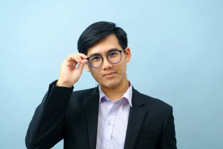 Téléchargez les photos : Portrait de jeune homme d'affaires asiatique intelligent portant des lunettes et un costume debout, souriant et regardant la caméra avec un fond isolé bleu clair. Concept d'entreprise. - en image libre de droit
