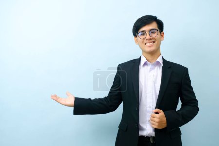 Photo for Portriat of asian young smart happy businessman dressed in suit standing straight, smiling, and pointing finger looking at camera with isolated light blue background. Business concept. - Royalty Free Image