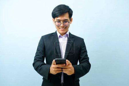 Foto de Portrait of young smart asian businessman standing and smiling while using smartphone with light blue isolated background. Business, connection concept. - Imagen libre de derechos